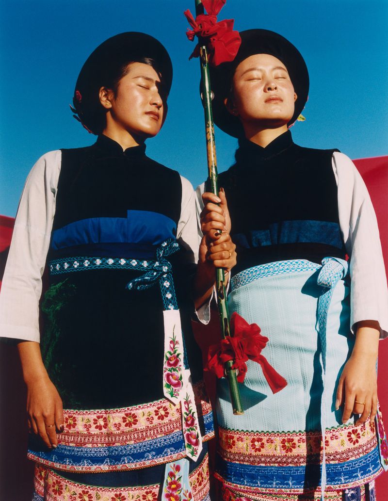 Two adult females in traditional clothing pose outdoors with a decorated bamboo pole against a vibrant blue and red backdrop.