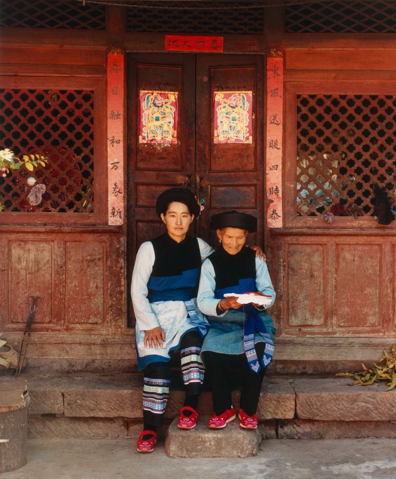Two women in traditional clothing sit on stone steps in front of an ornate wooden door, framed by decorative panels and Chinese script.