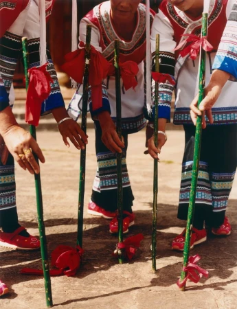 Four women in traditional clothing participate in a bamboo dance outdoors, holding green bamboo poles decorated with red fabric.