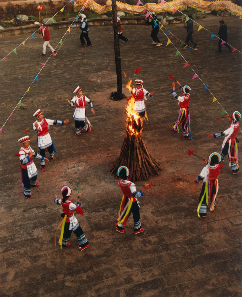 Group of people in traditional clothing dances in a circle around a bonfire in an outdoor, daytime performance setting.