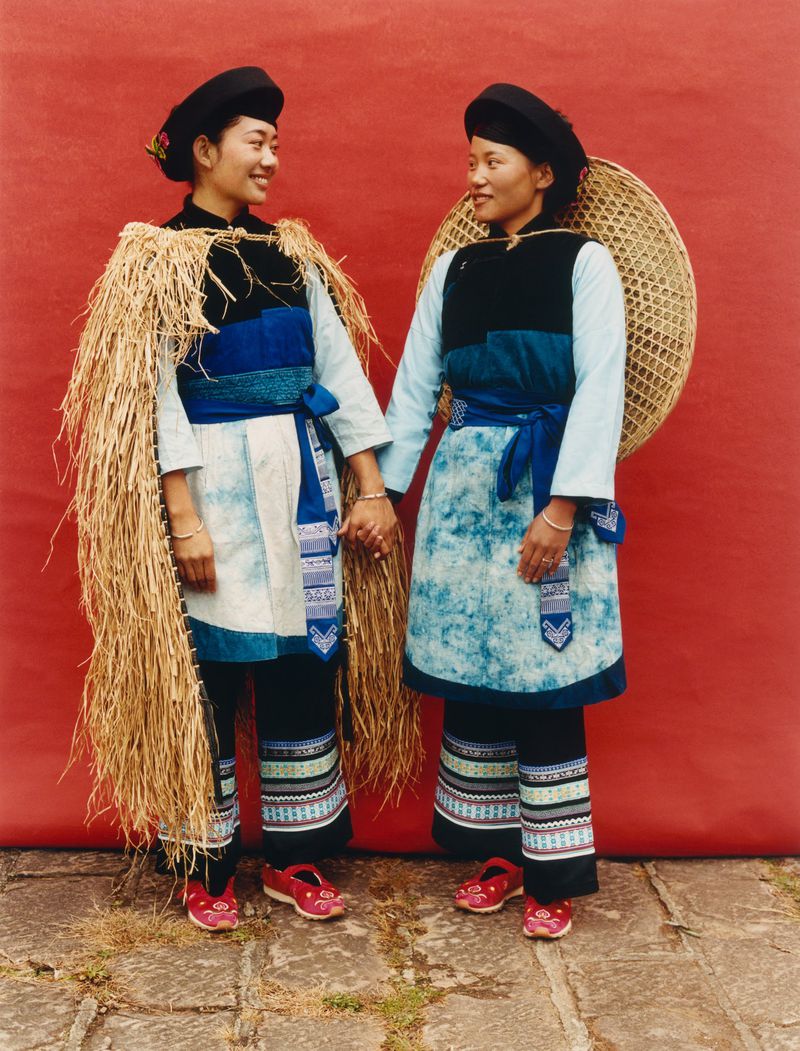 Two women in traditional clothing stand in front of a red wall, holding hands and smiling, with one wearing a straw cape.