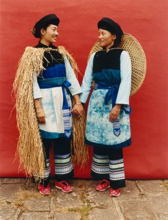 Two women in traditional clothing stand in front of a red wall, holding hands and smiling, with one wearing a straw cape.
