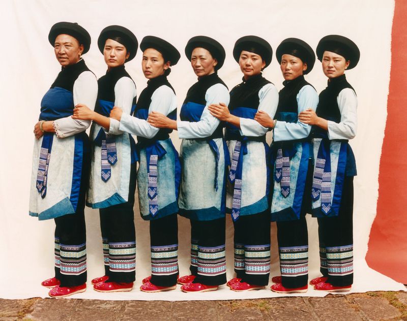 Seven Asian women stand in a row, wearing traditional blue and black outfits with patterned leg coverings and red shoes against white and red.