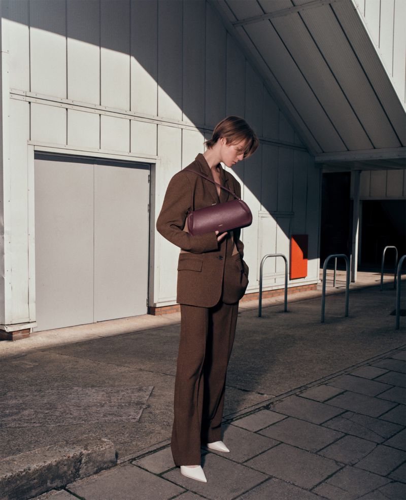 Adult woman wearing brown suit with a purple leather handbag stands on a concrete sidewalk in front of a white building.