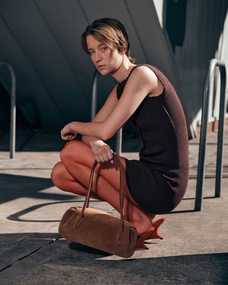 Stylish woman in a brown dress and heels poses crouching on concrete with a brown suede bag, under the light of a city day.