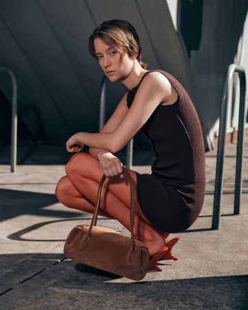 Stylish woman in a brown dress and heels poses crouching on concrete with a brown suede bag, under the light of a city day.