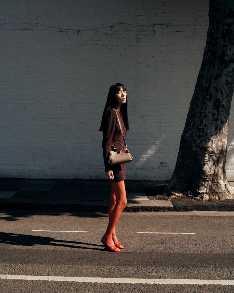 A long-haired woman in a brown dress poses with a beige handbag on a city street, illuminated by sunlight, alongside a large tree.