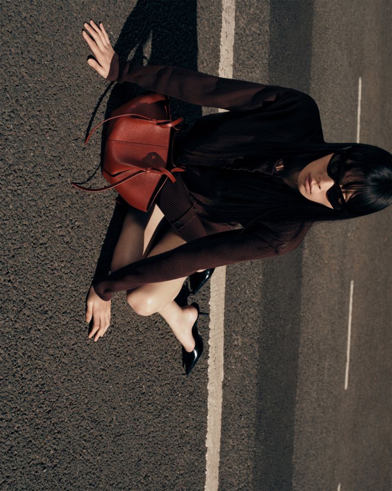Woman in brown outfit and sunglasses poses on asphalt street with red leather handbag and black heels in high-angle fashion shot.