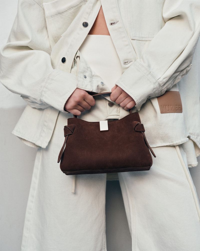 Woman in a light-toned denim jacket and pants holds a brown suede handbag, set against a neutral backdrop in a studio shot.