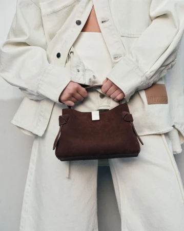 Woman in a light-toned denim jacket and pants holds a brown suede handbag, set against a neutral backdrop in a studio shot.