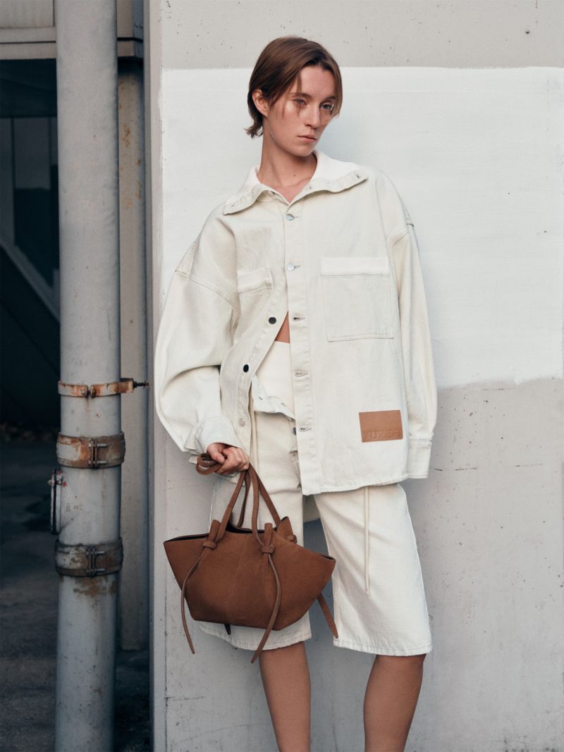 Woman in matching off-white jacket and shorts poses with a brown leather tote bag against an urban wall backdrop.