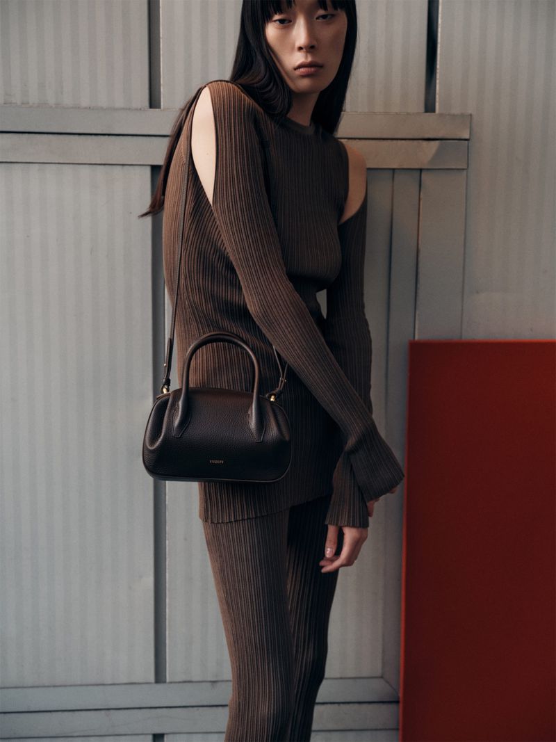 An Asian woman poses in a ribbed brown cut-out dress, holding a brown leather handbag in an indoor setting with gray and red accents.
