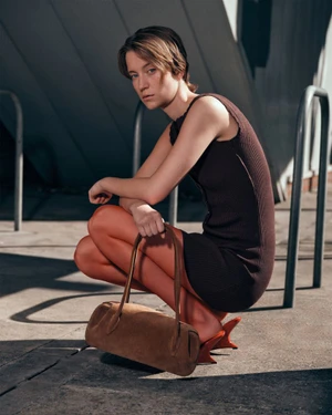 Model in brown dress crouching and holding a brown suede handbag outdoors in an urban setting