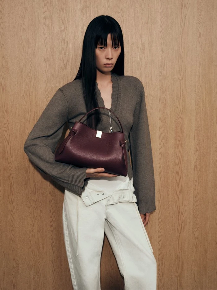 Model holding a dark burgundy leather handbag, wearing a grey knit top and white trousers against a wood grain wall.