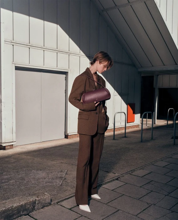 Model in a brown suit holding a cylindrical burgundy handbag standing outdoors in harsh sunlight