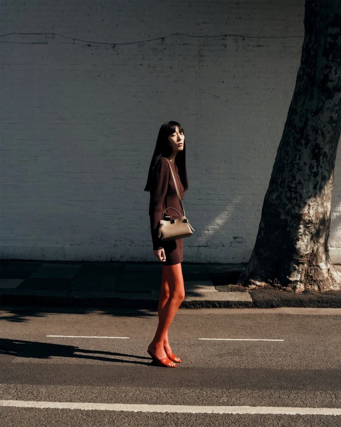 Model wearing a dark brown dress and shoulder bag standing on an urban street beside a large tree trunk against a white brick wall.