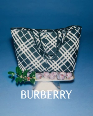 Burberry plaid check tote bag on a silver pedestal tray with plums and holly leaves against a blue background.