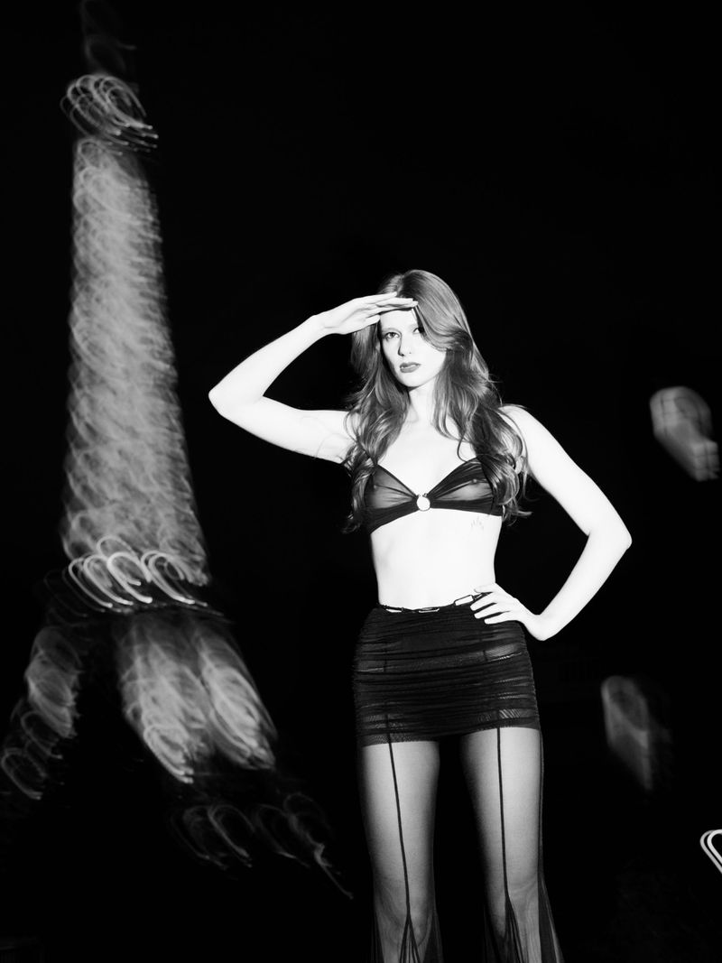 Monochrome fashion portrait of a woman in lingerie with a hand shielding her eyes, posing in front of blurred lights.