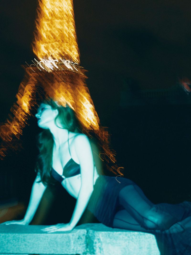 Woman in bra and skirt poses at night in blurred motion with the Eiffel Tower visible in the background, creating an editorial style image.