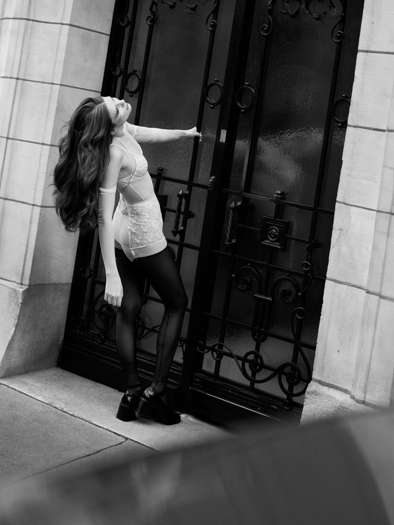 A woman with long hair, wearing lingerie and heels, leans against an ornate black door in a monochromatic outdoor fashion shot.