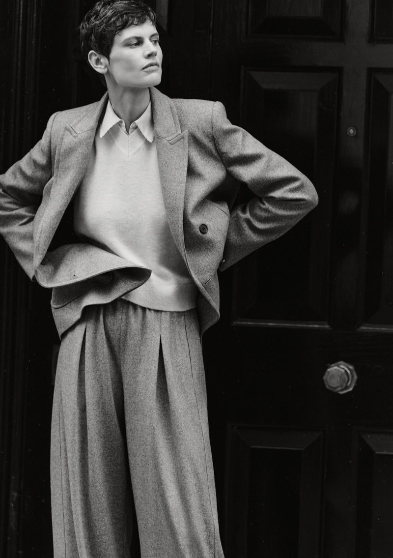 Monochrome shot of a woman in a grey suit with a white shirt, hands on hips, posing in front of a black paneled door, looking away.