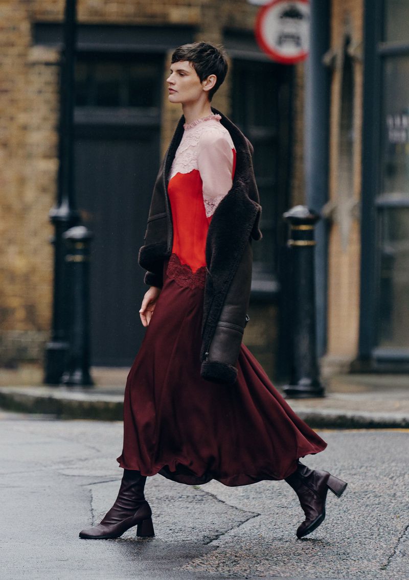 Adult woman in stylish red dress and brown jacket walks confidently on a city street with brick buildings in the background on an overcast day.