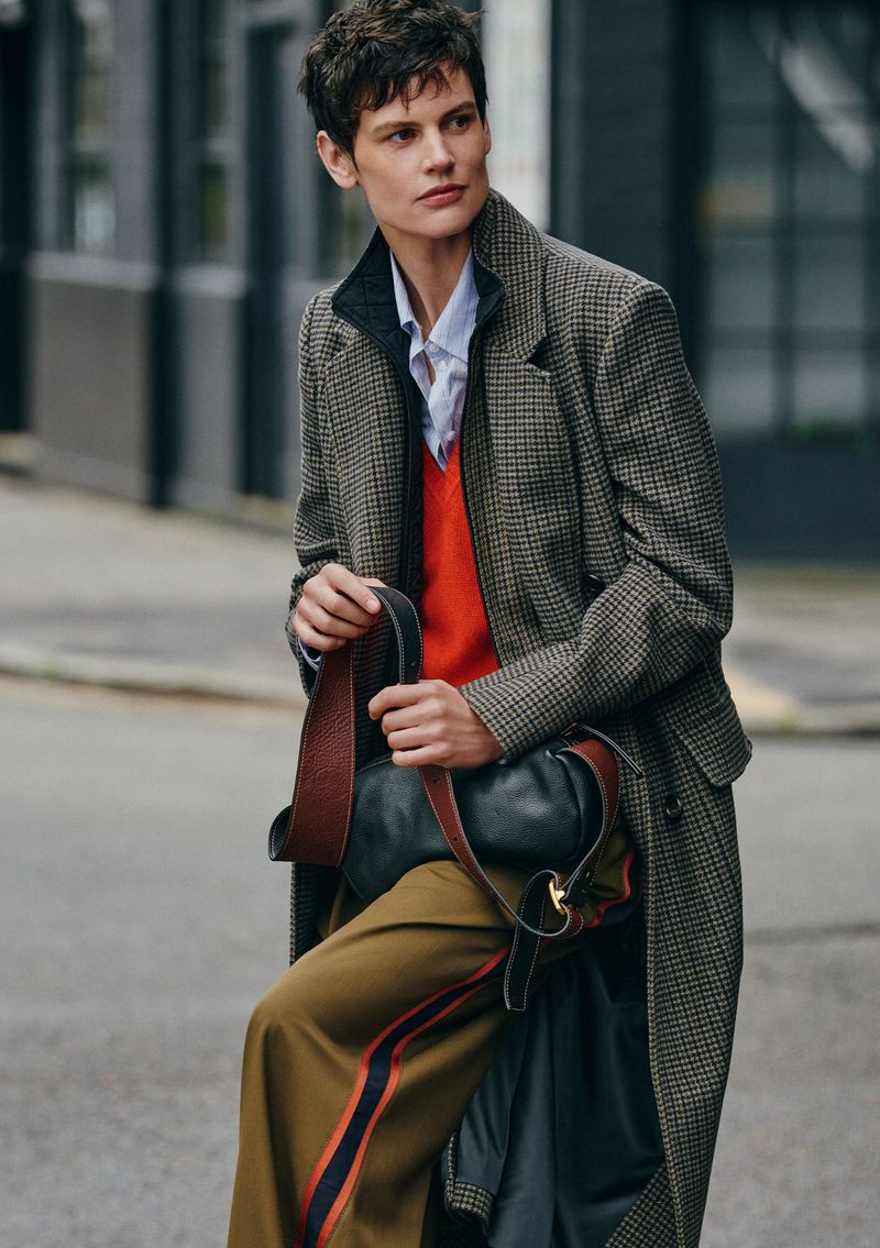 A woman in a tweed coat and orange sweater walks on a city street, holding a black purse with a shoulder strap. Autumn fashion.