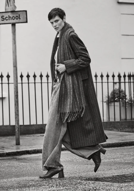 Monochrome portrait of an adult female with short hair walking in the street wearing coat, scarf, denim pants, and boots.