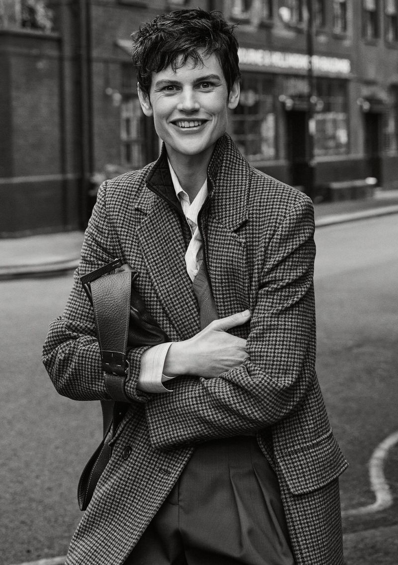 Monochrome portrait of smiling adult woman with short hair wearing a patterned blazer and carrying a dark leather bag on a city street.