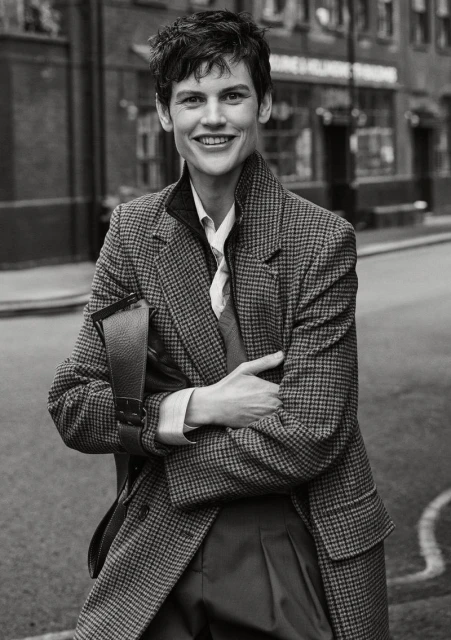 Monochrome portrait of smiling adult woman with short hair wearing a patterned blazer and carrying a dark leather bag on a city street.