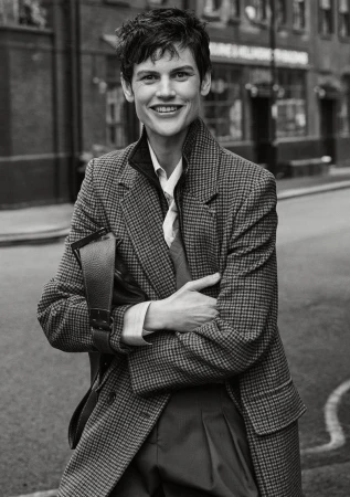 Monochrome portrait of smiling adult woman with short hair wearing a patterned blazer and carrying a dark leather bag on a city street.