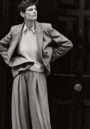 Model Saskia de Brauw wearing a tailored grey suit and white collared shirt, posing against a dark paneled door.