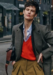 Model Saskia de Brauw in a houndstooth coat, red sweater, and pleated khaki trousers on an urban street.