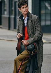 Saskia de Brauw wearing a checked tweed coat, red knitwear, and olive trousers holding a leather handbag on a city street.