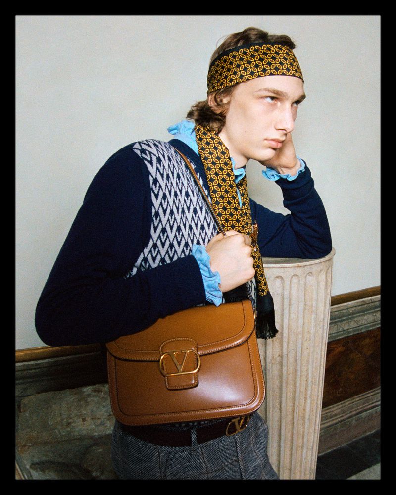 A young adult male model poses with a leather satchel, wearing a patterned headband and sweater, leaning against a marble column indoors.