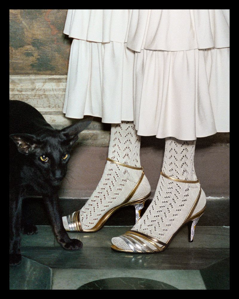 Fashion shot featuring two figures in white layered dresses and patterned socks with metallic heels, accompanied by a sleek black cat.
