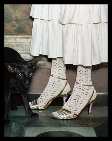 Fashion shot featuring two figures in white layered dresses and patterned socks with metallic heels, accompanied by a sleek black cat.