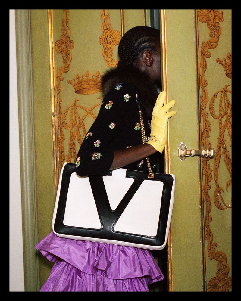 Woman in floral black cardigan, purple ruffle skirt, and yellow gloves with a white-and-black Valentino handbag stands by ornate doors.