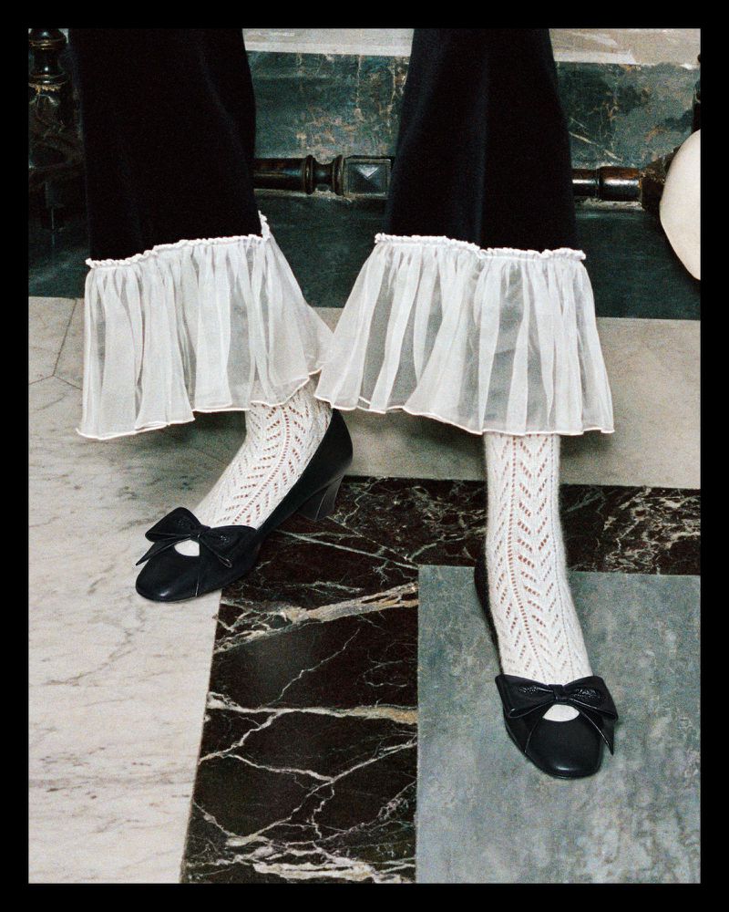 Close-up of black trousers with white frills, patterned white socks, and black bow-tie shoes on a marble floor, showcasing fashion.
