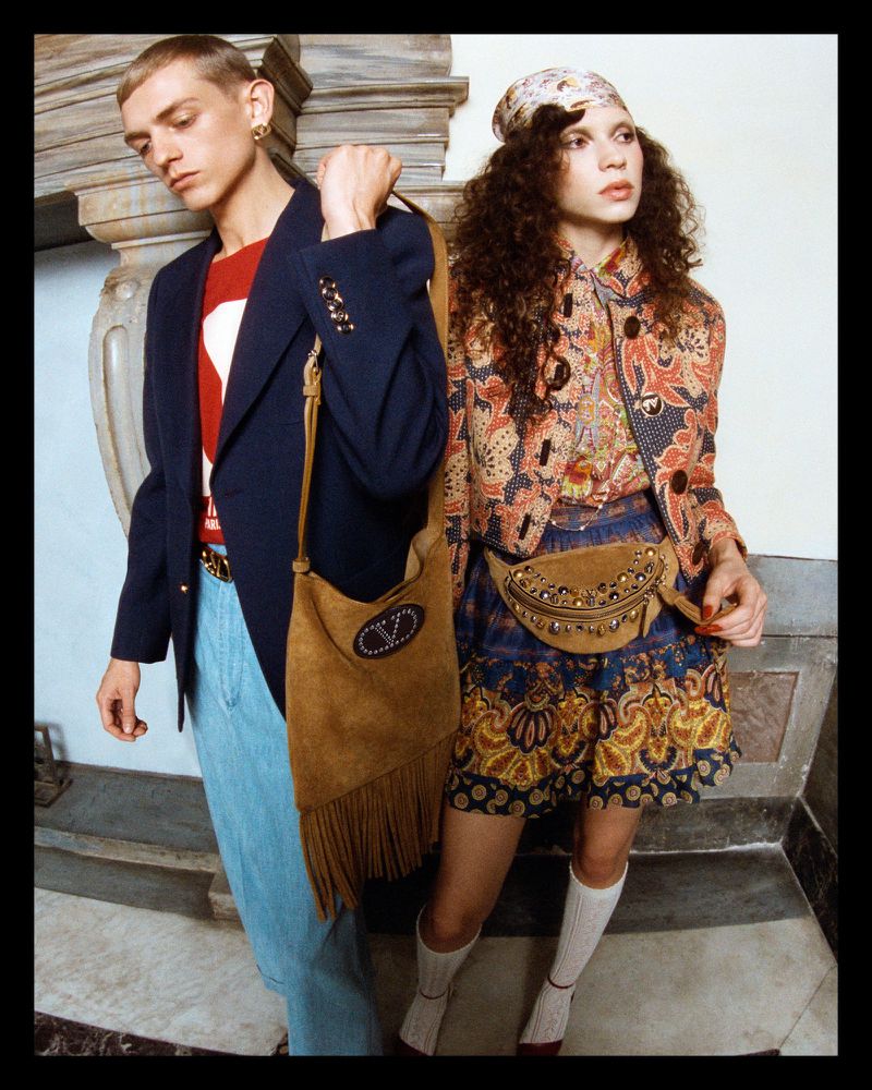 Two fashion models pose in patterned vintage-inspired clothing with suede handbags in an indoor studio with neutral tones. 