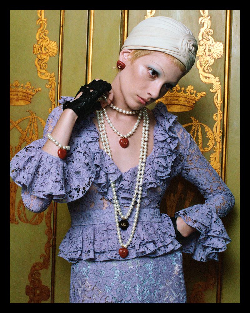 A woman poses wearing a lilac lace dress, pearl necklaces, and a white turban, against a decorative gold and green patterned background.