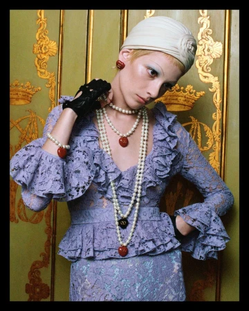 A woman poses wearing a lilac lace dress, pearl necklaces, and a white turban, against a decorative gold and green patterned background.