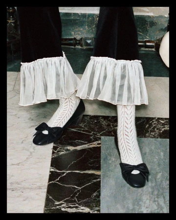 Close up of feet in white patterned socks and black bow pumps under dark trousers with white ruffled cuffs.