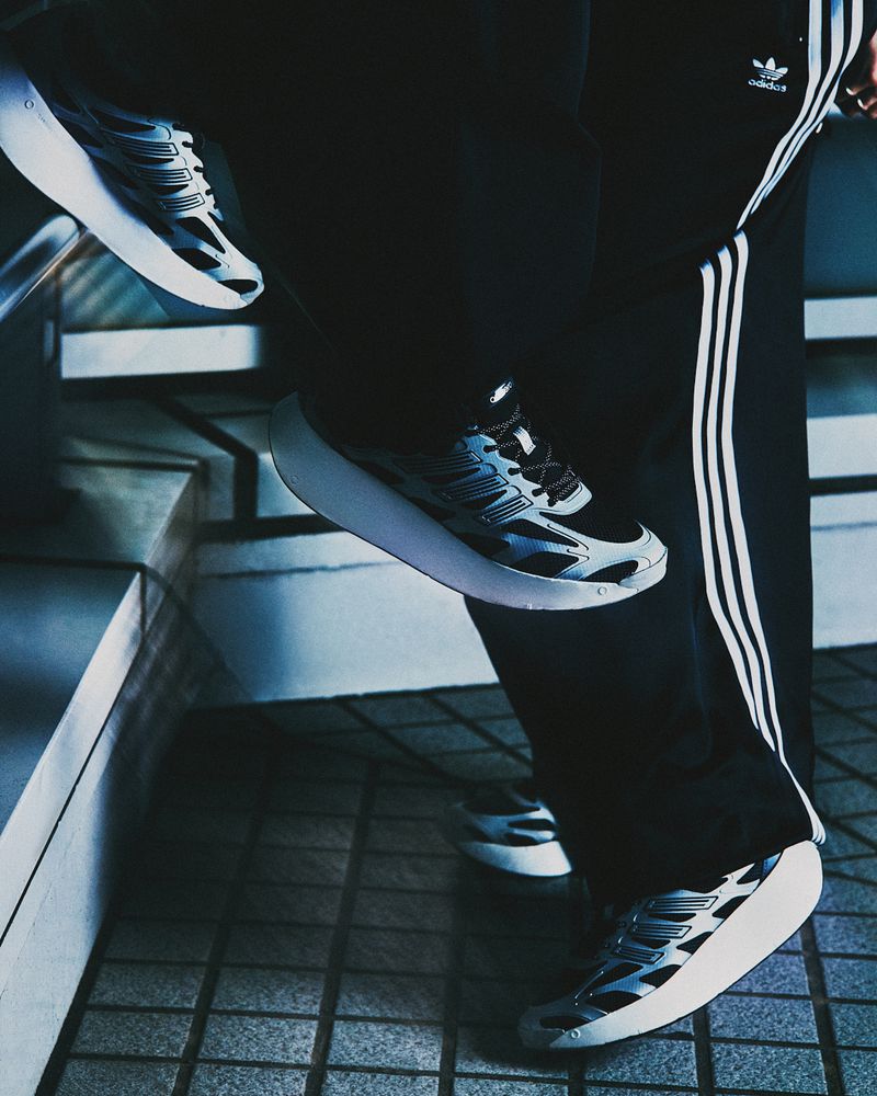 Three pairs of Adidas running shoes are positioned mid-air in front of a person wearing Adidas track pants with the brand's logo.