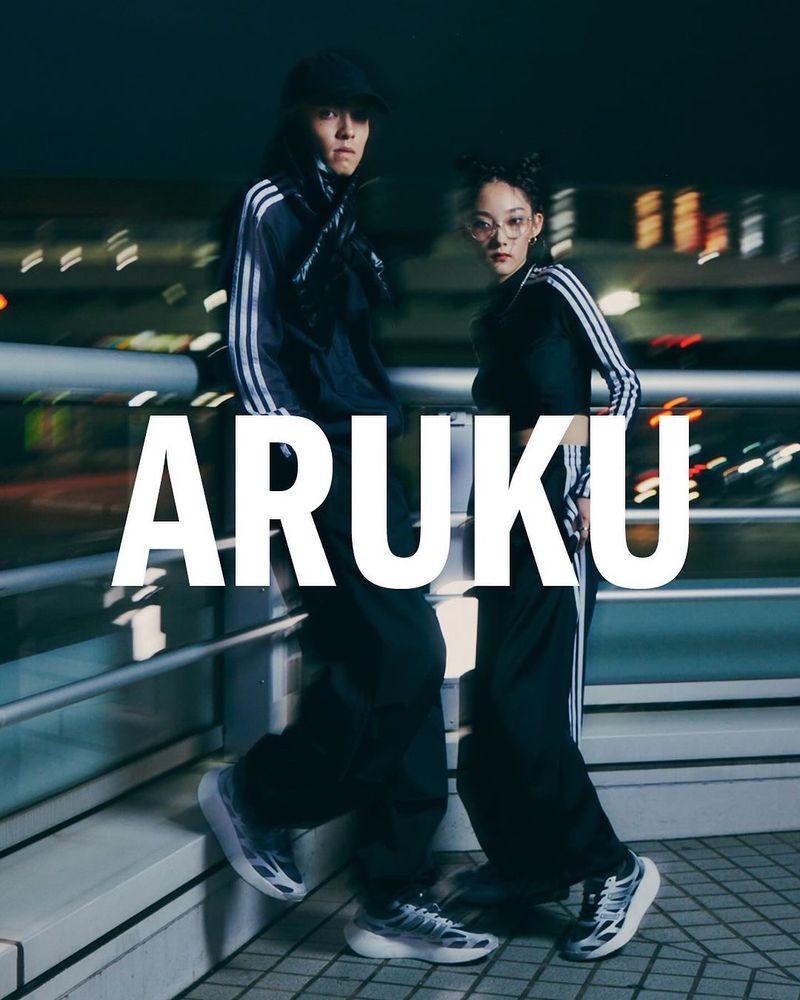Two stylish people in Adidas tracksuits and sneakers pose outdoors at night with blurred lights in the background, showcasing a sporty fashion aesthetic.