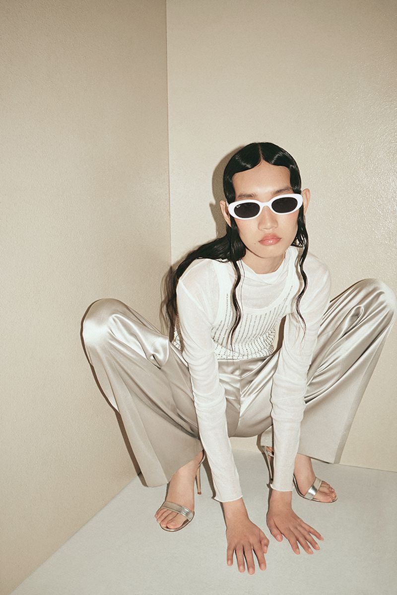 A slender model poses with dark hair and white sunglasses, wearing a white top and silver pants and heels, in a photo studio setting.