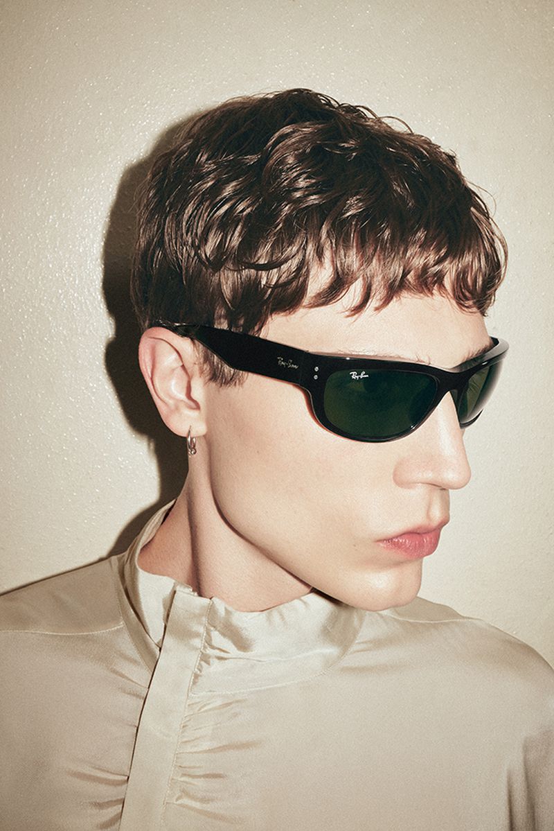Close-up of a young adult male model wearing black Ray-Ban sunglasses and a beige shirt, posing indoors against a neutral wall.