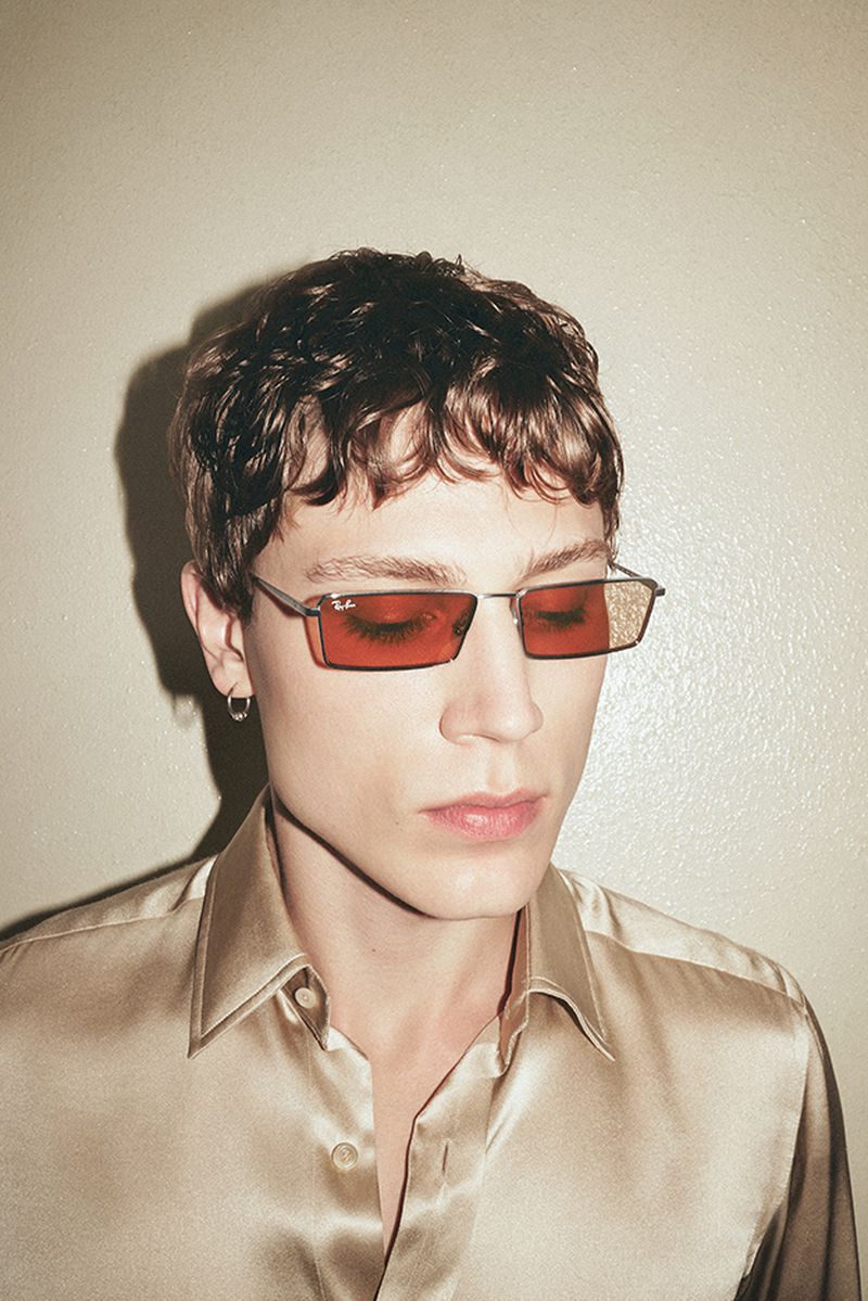 Portrait of a young adult male with curly hair wearing a gold satin shirt and red tinted sunglasses against a neutral background.
