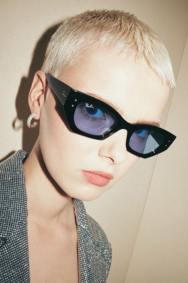 Close-up of a stylish woman with short blonde hair wearing black Ray-Ban sunglasses and a sparkling blazer in a studio setting.