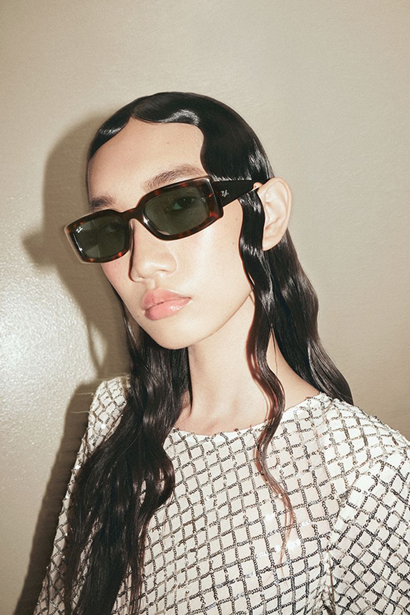 Close-up of a young adult wearing stylish sunglasses, posing indoors with slicked-back dark hair and a sequined white top.